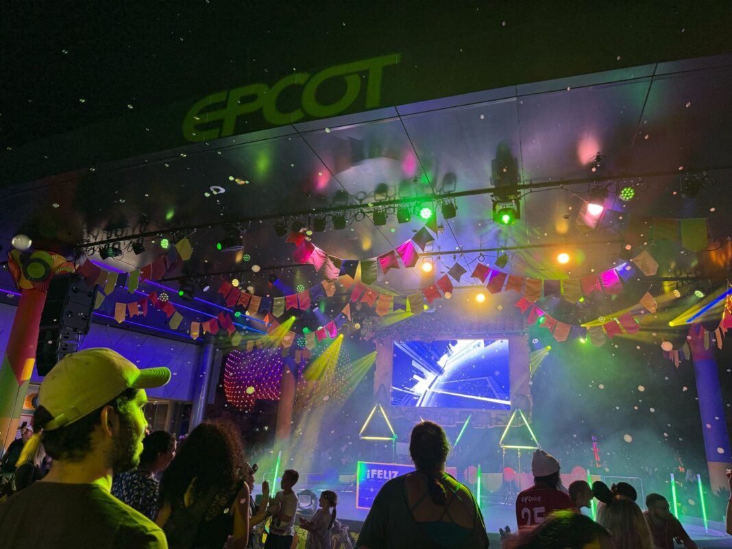 Amidst vibrant lights and geometric decorations, a DJ electrifies the stage at EPCOT, the screen proclaiming "¡Feliz Año Nuevo!" in celebration of New Year's Eve 2025. The pulsating beats set the perfect backdrop for an unforgettable dance party.
