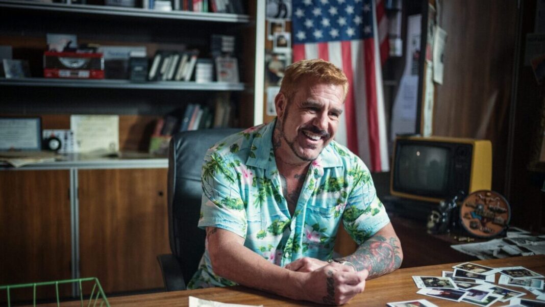 A white man in a tropical button-down is sitting at a desk.